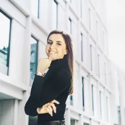 Uśmiechnięta kobieta w czarnym golfie i skórzanej spódnicy pozuje na tle nowoczesnego biurowca z dużymi oknami.