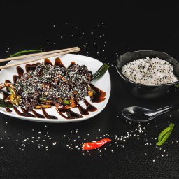Studio Migafka - Stylizowane danie azjatyckie z mięsem i warzywami polane sosem i posypane sezamem, podane z ryżem, pałeczkami i łyżką na czarnym tle z rozsypanym sezamem.