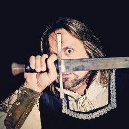 Studio Migafka - Mężczyzna w kostiumie z mieczem zasłaniającym oko, ciemne tło, skórzana zbroja na ramieniu, biała koszula z koronkowym wykończeniem.