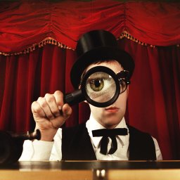 Studio Migafka - Mężczyzna w cylindrze i muszce, z powiększonym okiem przez lupę na tle czerwonej kurtyny, stylizowane ujęcie.