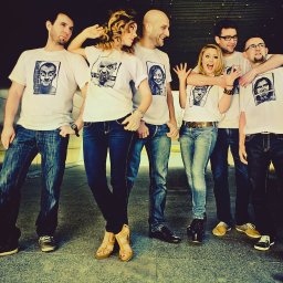 Studio Migafka - Grupa sześciu osób pozuje do zdjęcia w białych koszulkach z nadrukami portretów, stojąc na szarym tle z ciemnymi płytkami.