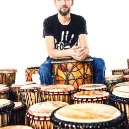 Studio Migafka - Młody mężczyzna z brodą siedzi na bębnie djembe, otoczony innymi bębnami, ubrany w czarny t-shirt z białym nadrukiem dłoni.