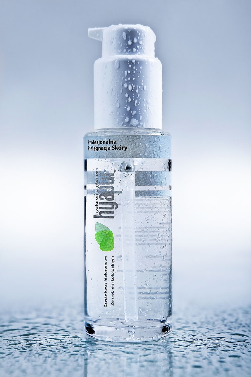 Butelka serum hialuronowego 'hyapur intense' z kroplami wody na powierzchni, umieszczona na mokrej, błyszczącej powierzchni, w tle rozmyte, jasne tło.