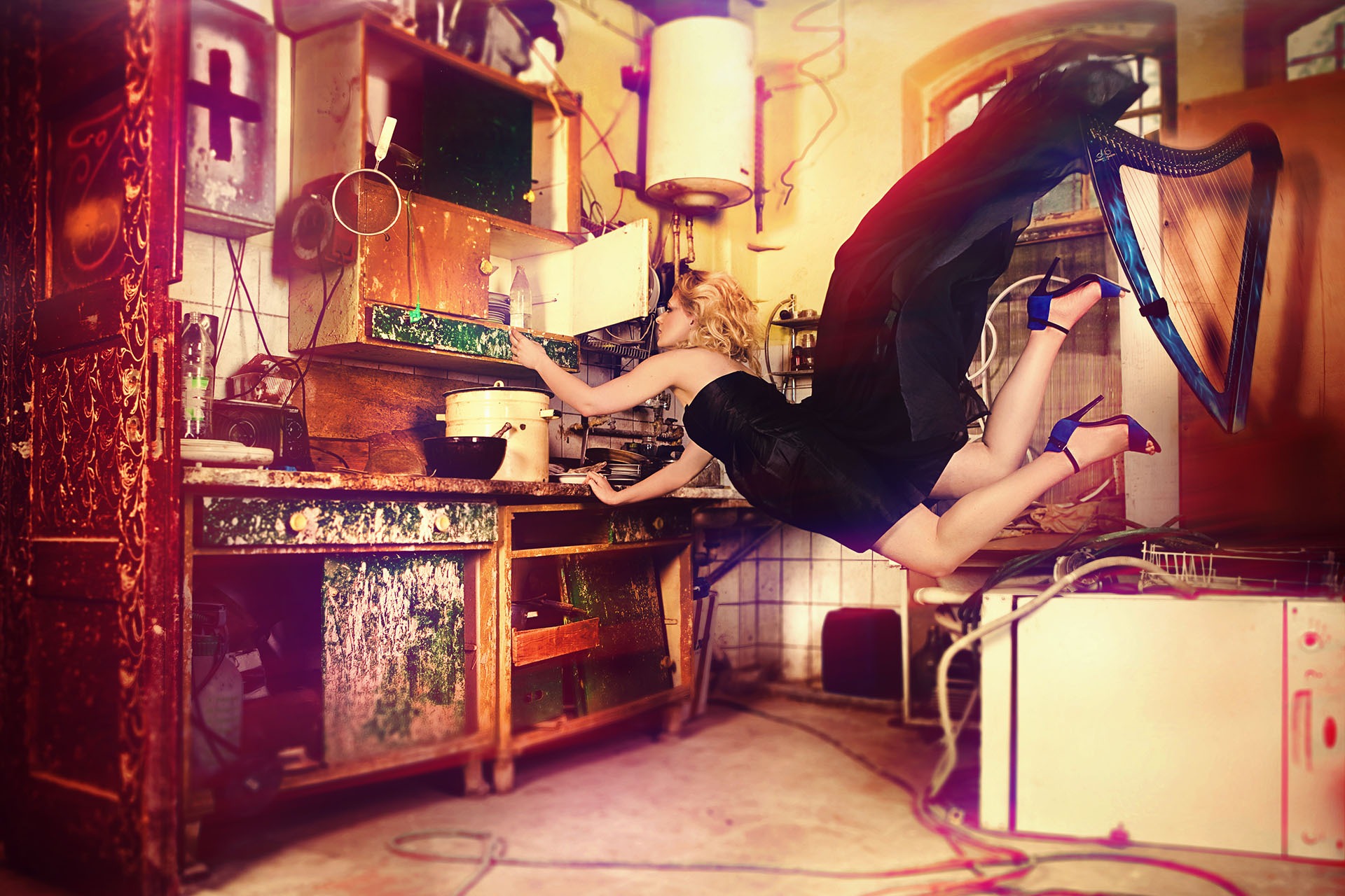 Kobieta w czarnej sukience i niebieskich szpilkach unosi się w powietrzu w kuchni, sięgając ręką w stronę szafki, z harfą obok. Scena surrealistyczna, oświetlona w sposób teatralny.