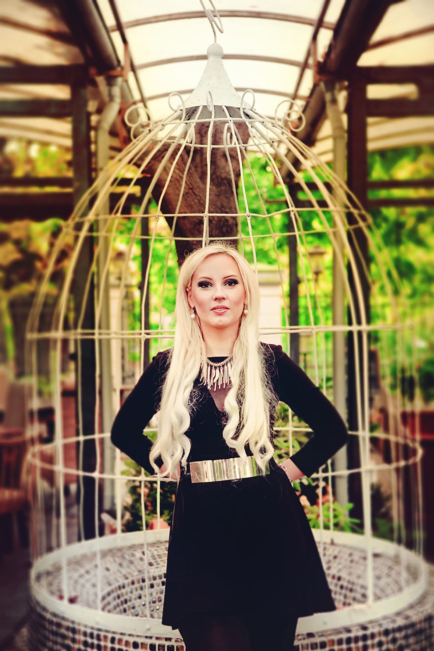 Portret kobiety we Wrocławiu, w czarnej sukience z szerokim złotym paskiem i ozdobnym naszyjnikiem, pozującej na tle białej dekoracyjnej klatki i zieleni.