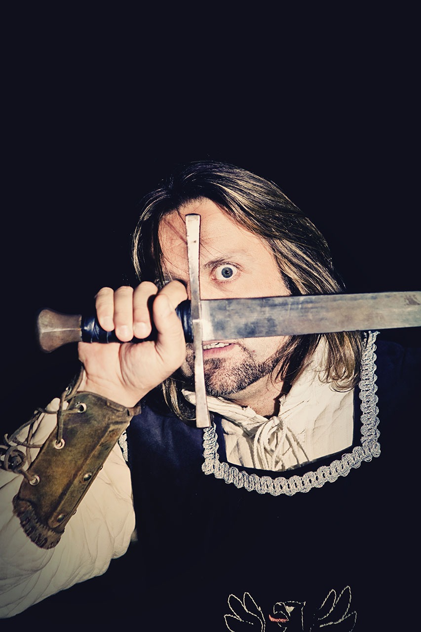 Mężczyzna w kostiumie z mieczem zasłaniającym oko, ciemne tło, skórzana zbroja na ramieniu, biała koszula z koronkowym wykończeniem.