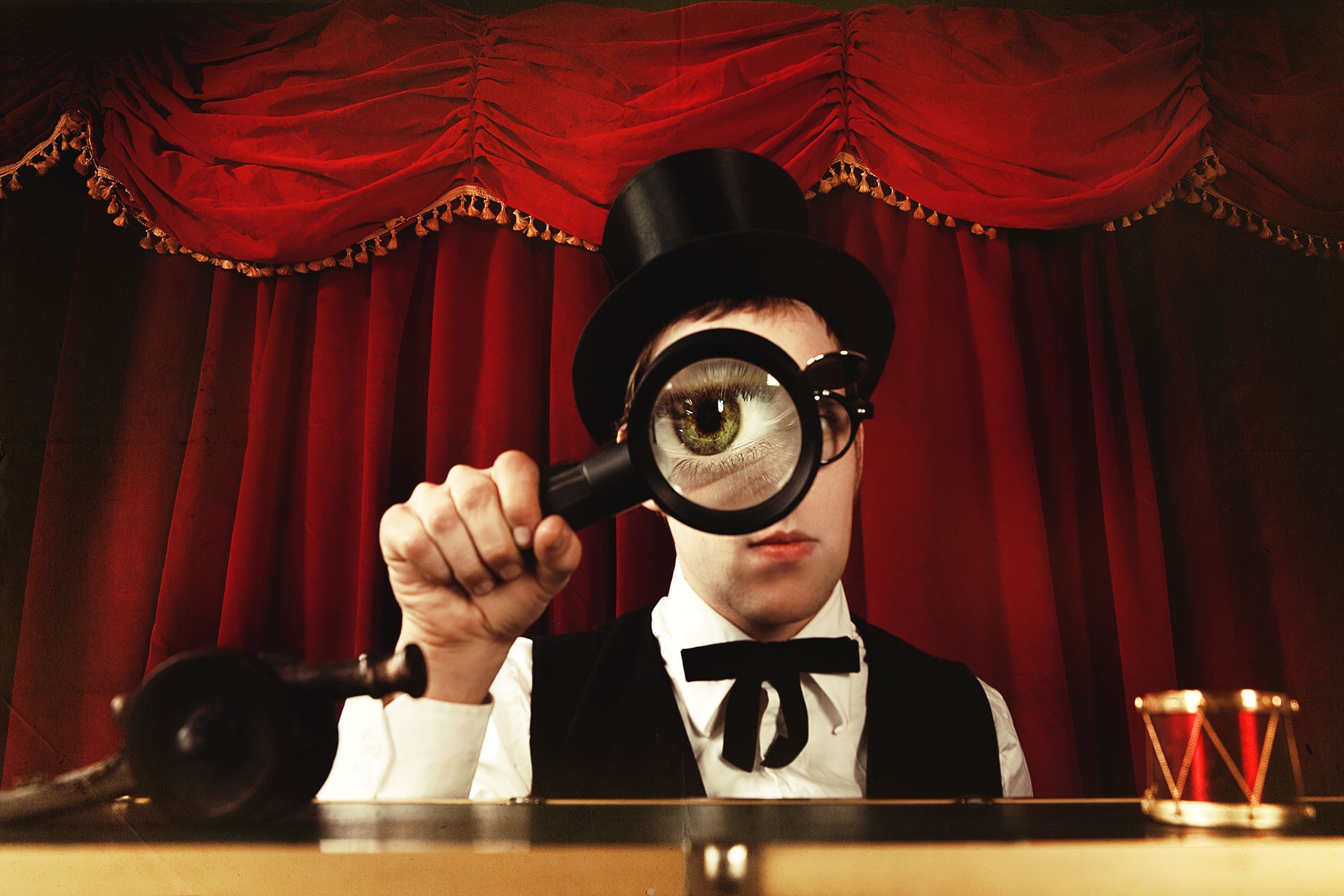 Mężczyzna w cylindrze i muszce, z powiększonym okiem przez lupę na tle czerwonej kurtyny, stylizowane ujęcie.
