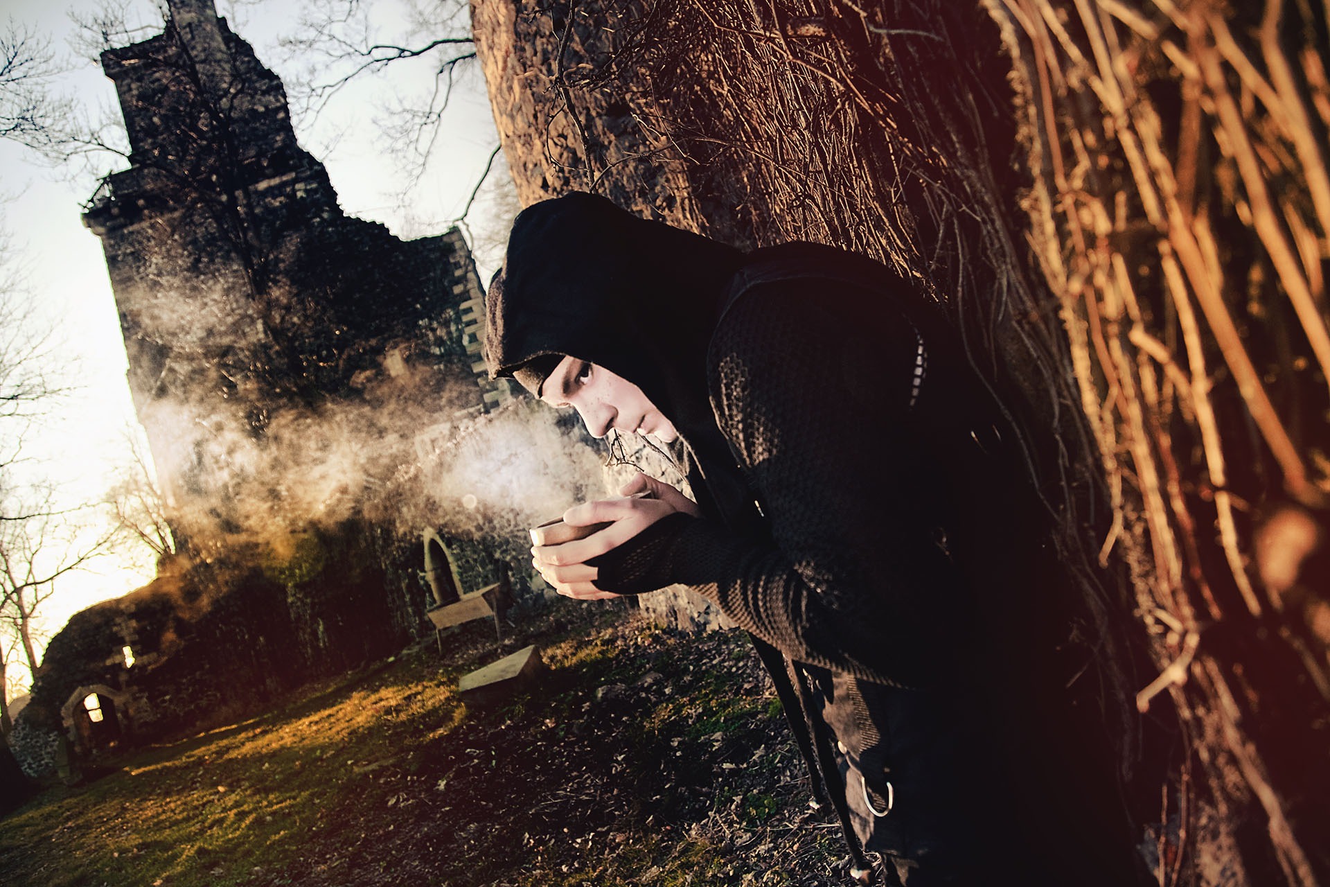 Młody mężczyzna w kapturze, stojąc przy drzewie z ruinami zamku w tle, dmucha parę ze złożonych dłoni w chłodny dzień.