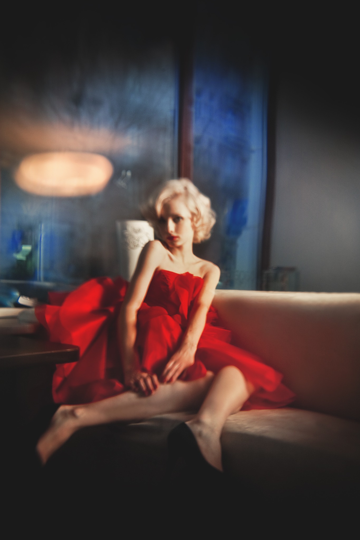 Kobieta o blond włosach i czerwonych ustach, ubrana w czerwoną suknię, siedzi na beżowej kanapie na tle okna z niebieskim światłem.