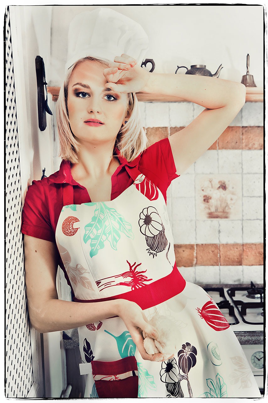 Kobieta w czapce kucharskiej i fartuchu z motywami roślinnymi opiera się o ścianę, trzymając w dłoniach ciasto na tle kuchni z kuchenką i imbrykiem.