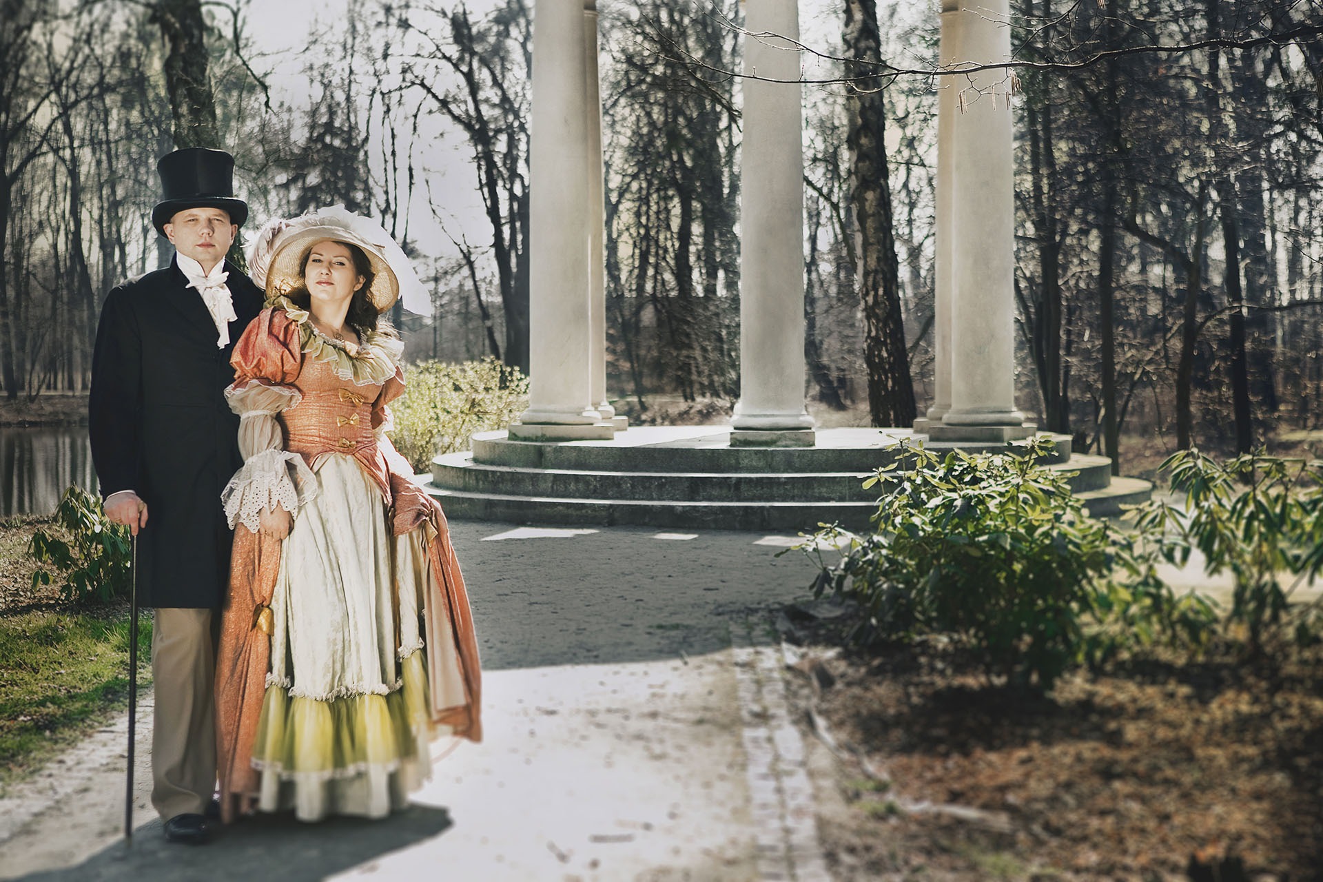 Para w strojach z epoki, on w cylindrze i fraku, ona w bogato zdobionej sukni i kapeluszu, pozują na tle kolumn i parku.