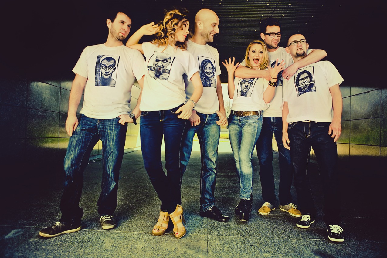 Grupa sześciu osób pozuje do zdjęcia w białych koszulkach z nadrukami portretów, stojąc na szarym tle z ciemnymi płytkami.