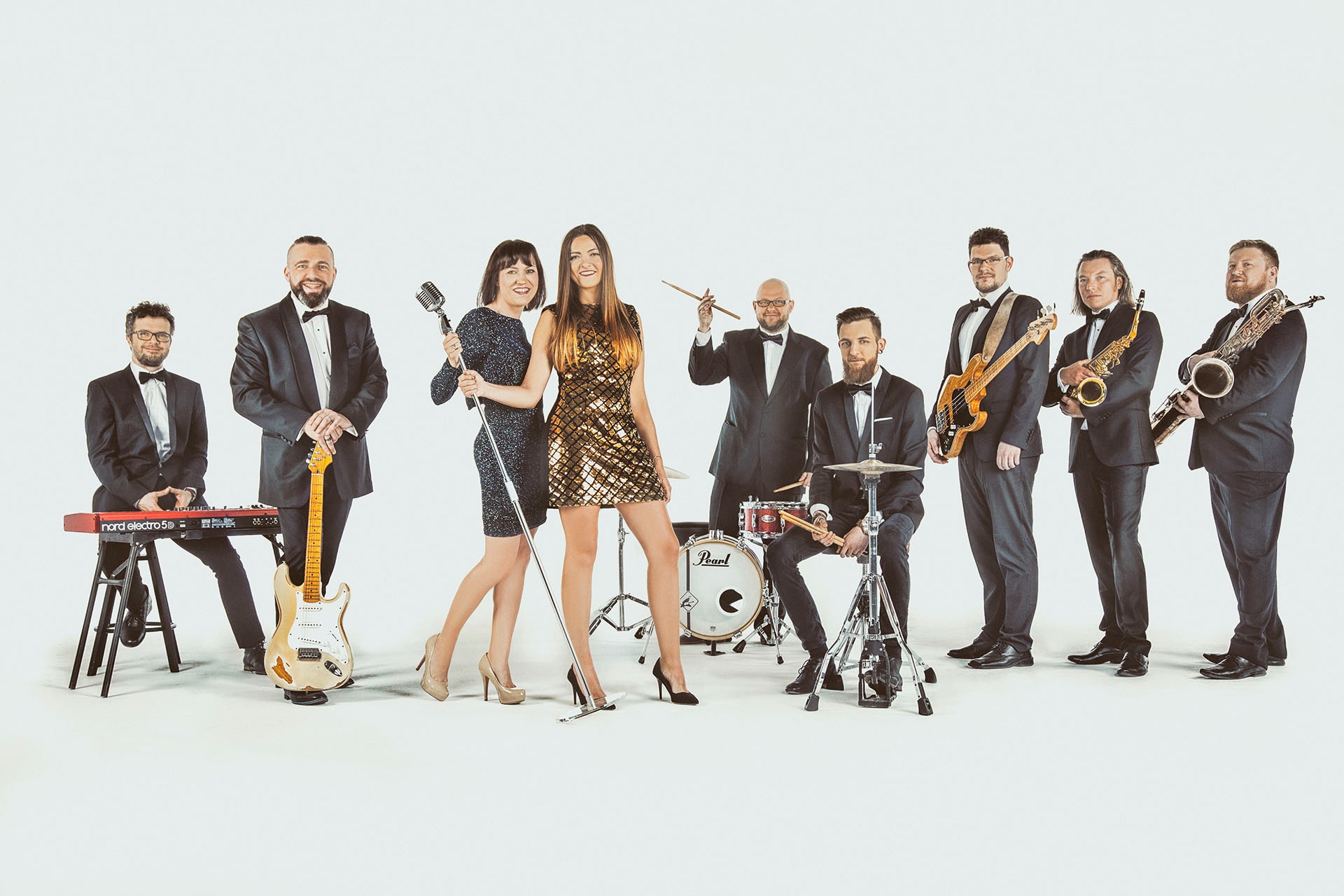 Zespół muzyczny w smokingach i sukienkach, z instrumentami (keyboard, gitara, perkusja, saksofon) pozuje na białym tle, kobieta w złotej sukience stoi na pierwszym planie.