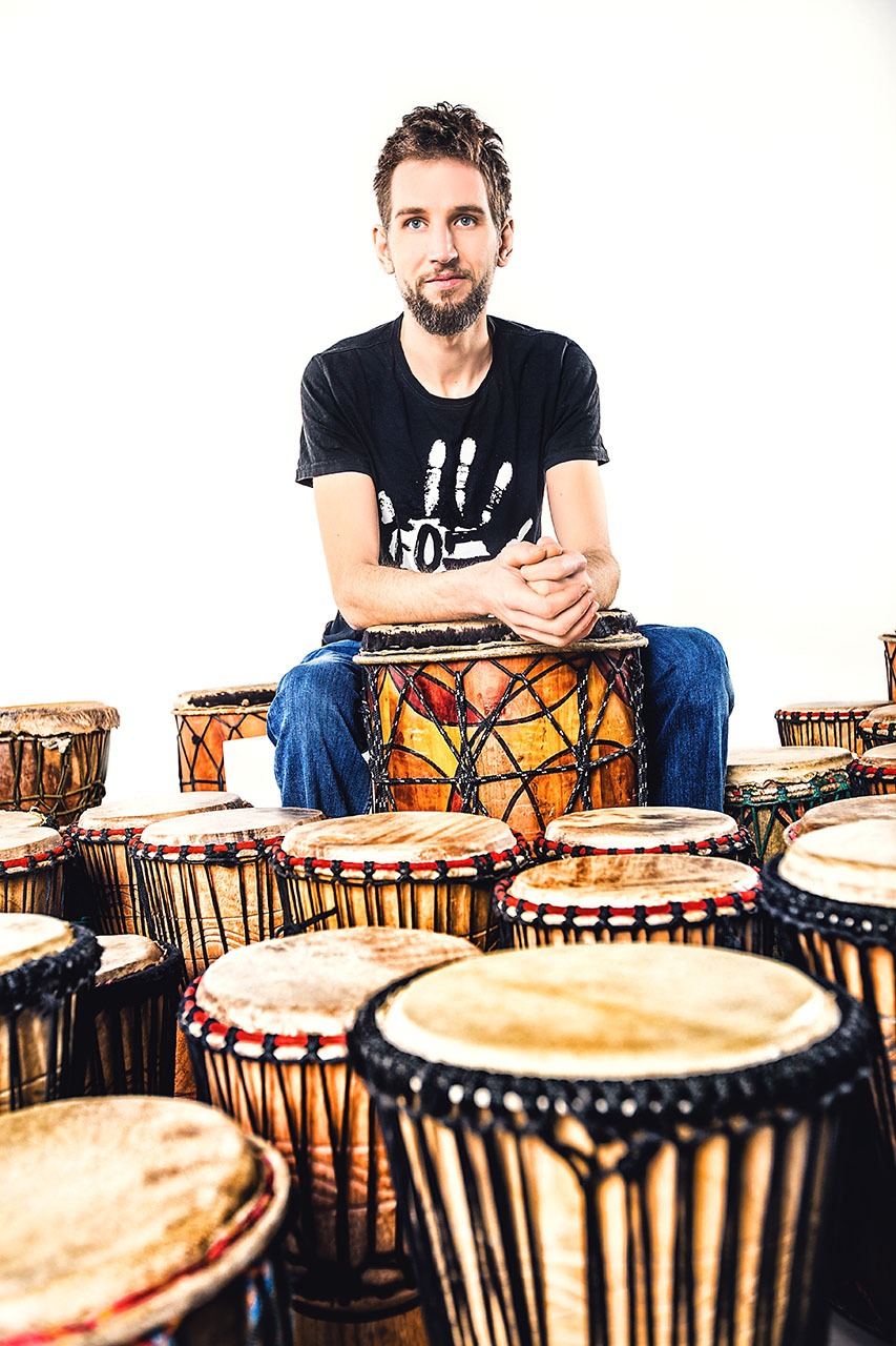 Młody mężczyzna z brodą siedzi na bębnie djembe, otoczony innymi bębnami, ubrany w czarny t-shirt z białym nadrukiem dłoni.