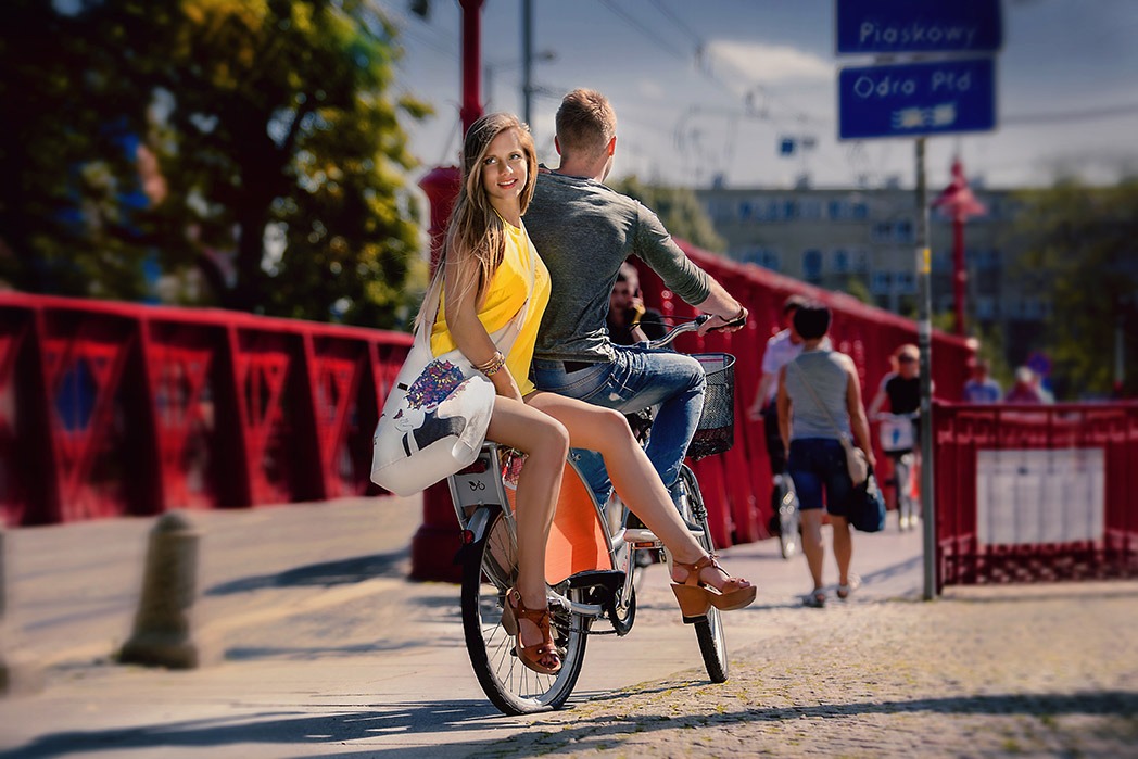 Para na rowerze miejskim przejeżdża przez czerwony most, kobieta w żółtej bluzce i brązowych sandałach odwraca się uśmiechnięta, trzymając torbę z ilustracją.