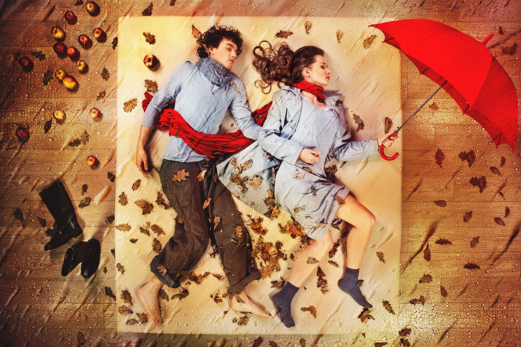 Para leżąca na beżowym materiale, otoczona jabłkami i jesiennymi liśćmi, kobieta trzyma czerwoną parasolkę, całość sfotografowana z góry, stylizowana na jesienną sesję.