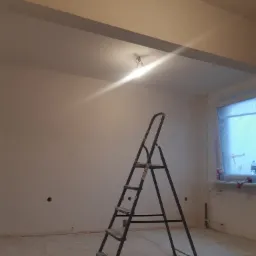 Pokój w trakcie malowania: ściany pokryte świeżą warstwą farby, drabina aluminiowa oparta o ścianę, okno zabezpieczone folią malarską, widoczne ślady po gładzi na podłodze.