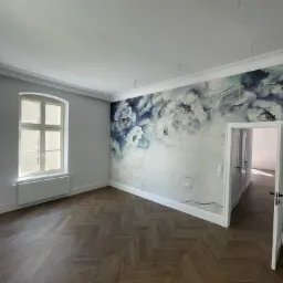 Jasne wnętrze z parkietem w jodełkę, ściana z motywem kwiatowym, okno z białymi ramami i widocznymi przewodami elektrycznymi na suficie.