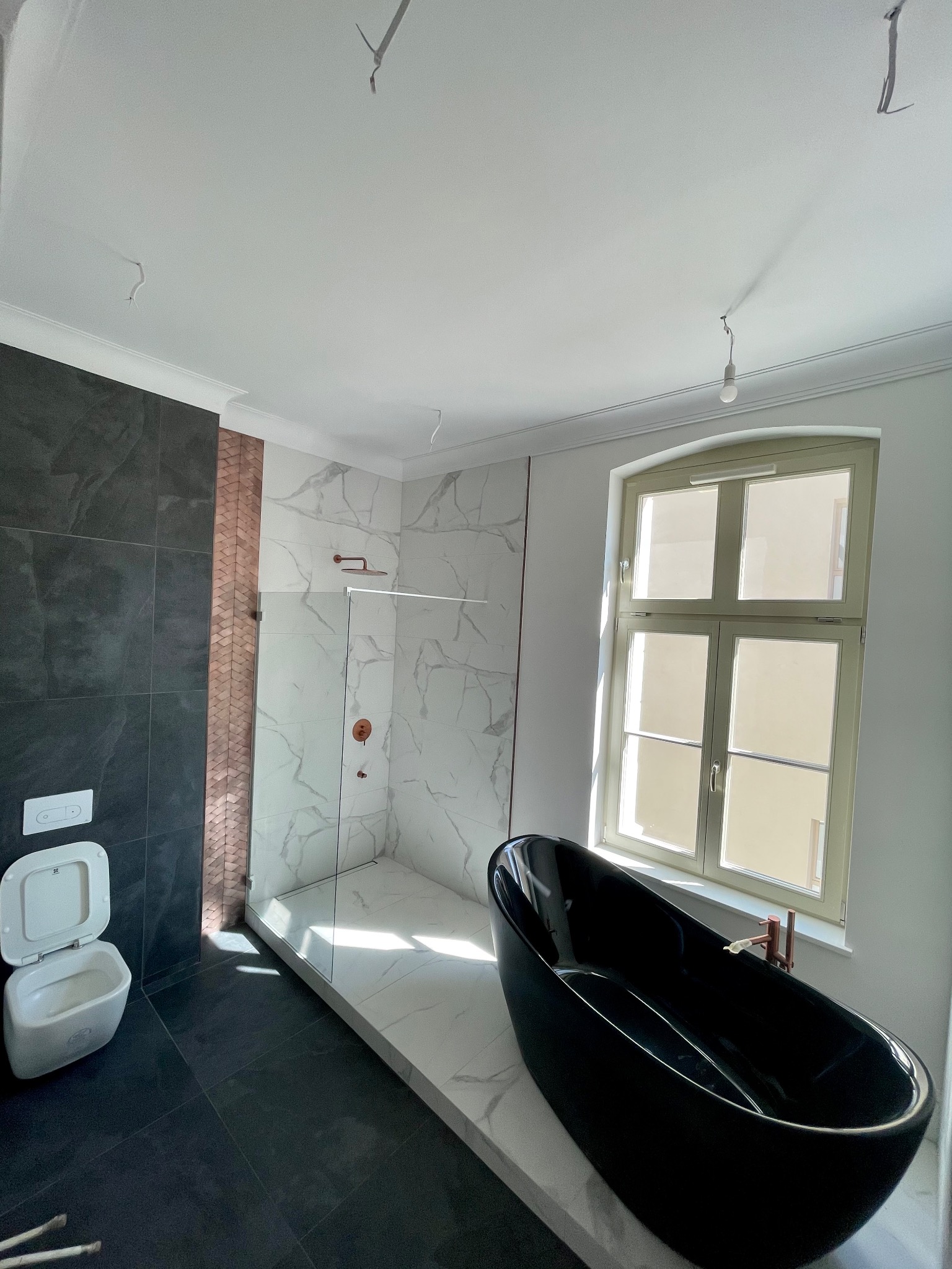 Nowoczesna łazienka w trakcie wykończenia: czarna wanna wolnostojąca, kabina prysznicowa walk-in z marmurowymi płytami, toaleta podwieszana i ciemne płytki podłogowe, widoczne niedokończone kable...