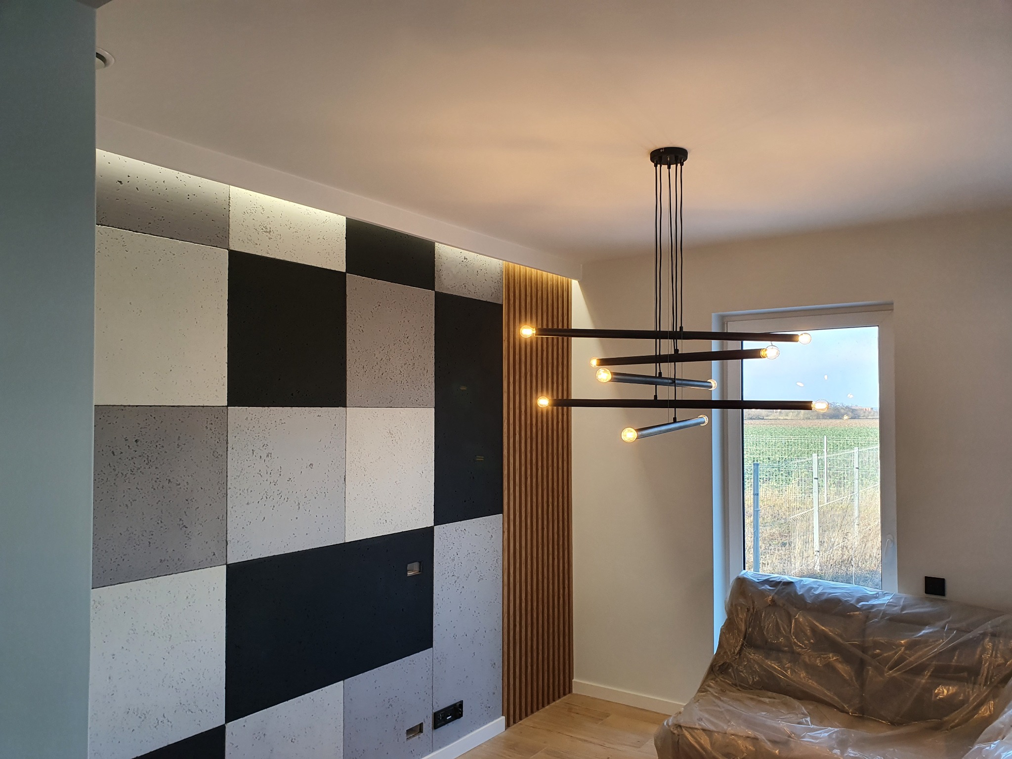 Nowoczesne wnętrze z dekoracyjną ścianą z paneli w odcieniach szarości i czerni, oświetlona designerską lampą wiszącą z żarówkami filamentowymi, z widokiem na zewnątrz przez okno.
