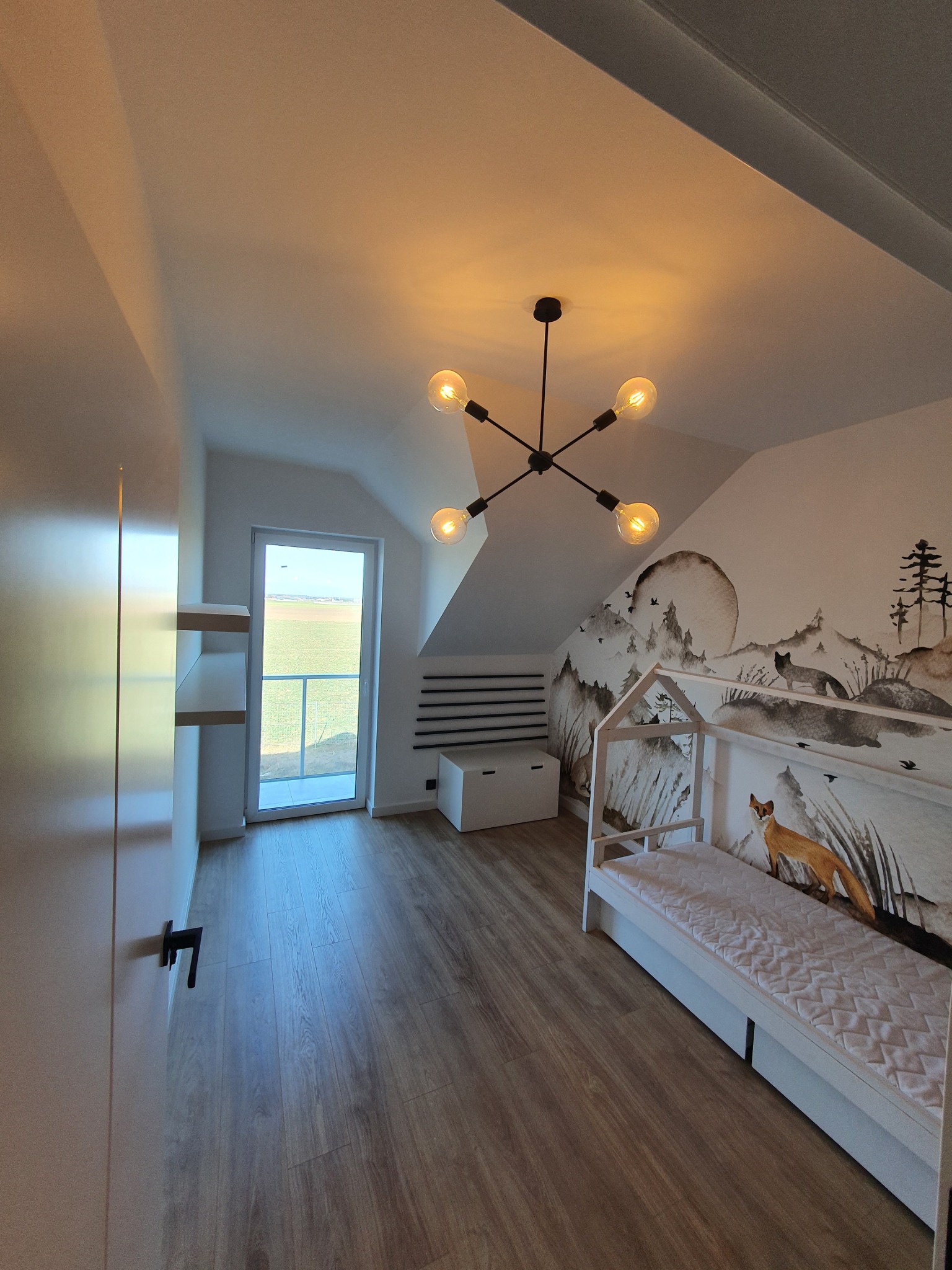 Pokój dziecięcy na poddaszu z łóżkiem w kształcie domku, ściennym malowidłem z motywem lasu i nowoczesną lampą wiszącą z żarówkami Edisona.