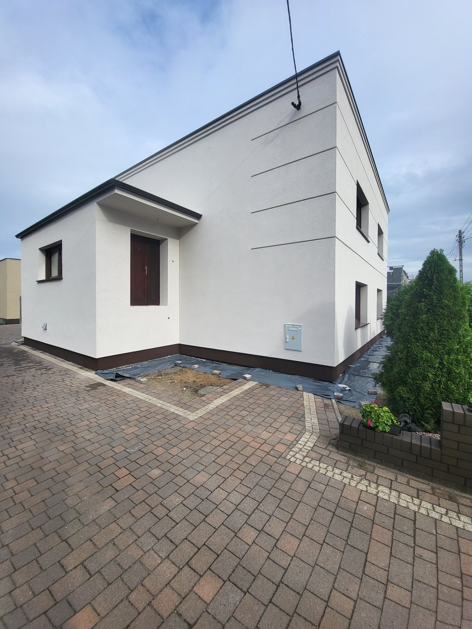Nowoczesny, jednopiętrowy dom z białą elewacją i brązowymi ramami okiennymi, widoczny z poziomu brukowanego podjazdu, z przygotowanym terenem pod nasadzenia wzdłuż fundamentów.