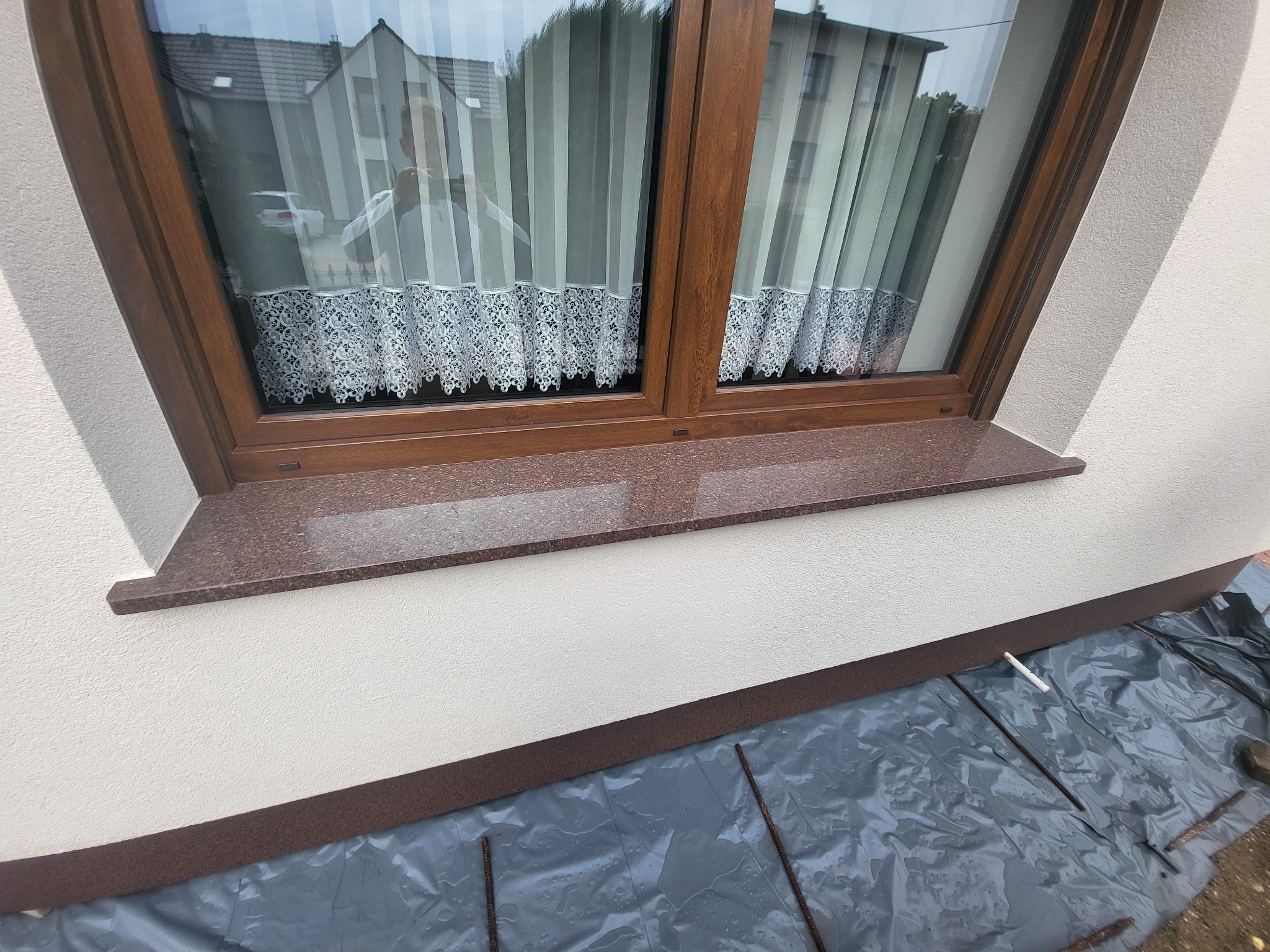 Zamontowany brązowy parapet z granitu na elewacji budynku, okno z firanką w tle, folia ochronna na ziemi.