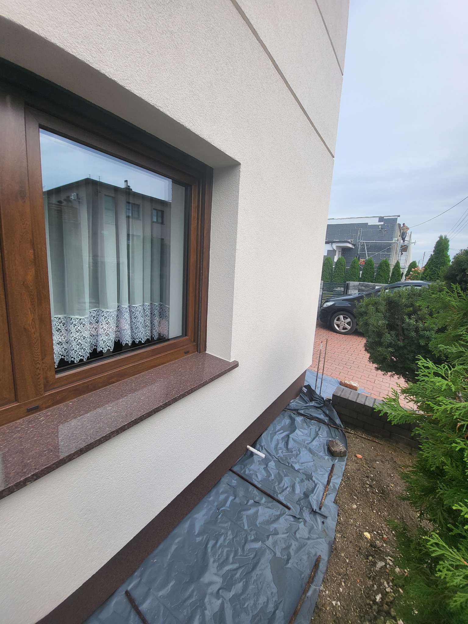 Elewacja budynku z oknem o brązowej ramie i marmurowym parapetem, zabezpieczona folią podczas prac na wysokości, widoczny pracownik na rusztowaniu i fragment ocieplonego budynku.