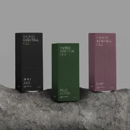 Trzy pudełka z olejkami eterycznymi: czarne z napisem Holy Basil, zielone z napisem Palo Santo i różowe z napisem Rose, ustawione na szarej skale. Pudełka mają minimalistyczny design...