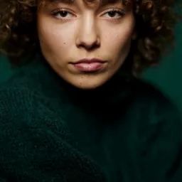 Portret kobiety o kręconych włosach i zielonym swetrze na jednolitym zielonym tle.
