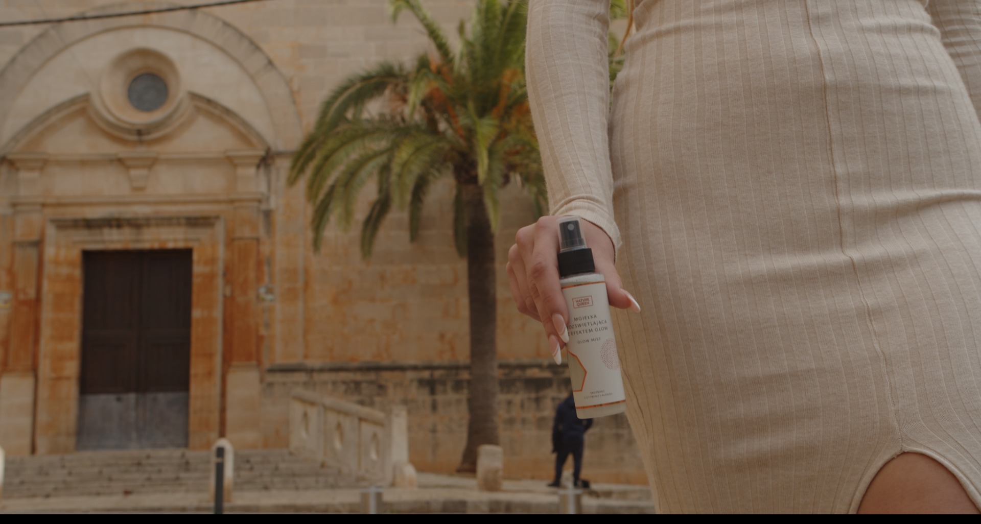 Kobieta w beżowej sukience trzymająca mgiełkę rozświetlającą do twarzy na tle kamiennej budowli i palmy.