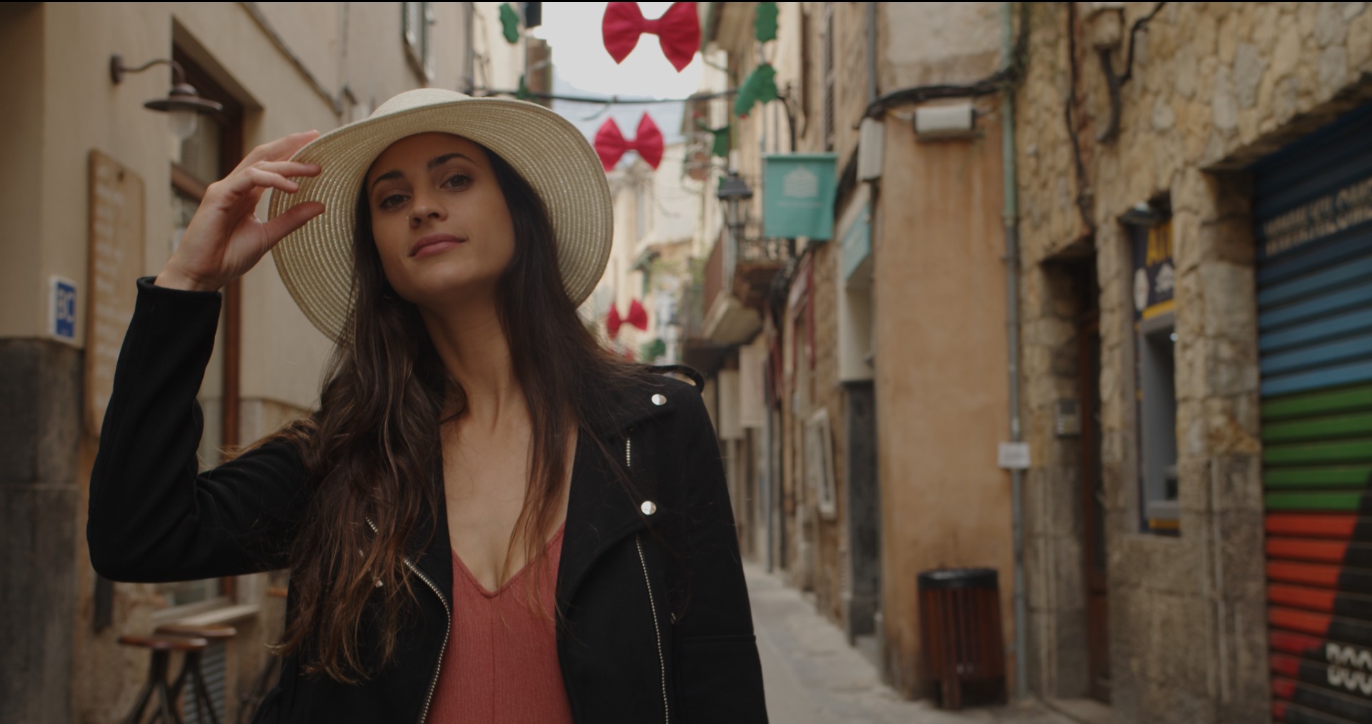 Kobieta w kapeluszu i czarnej kurtce pozuje na tle wąskiej uliczki z dekoracjami.