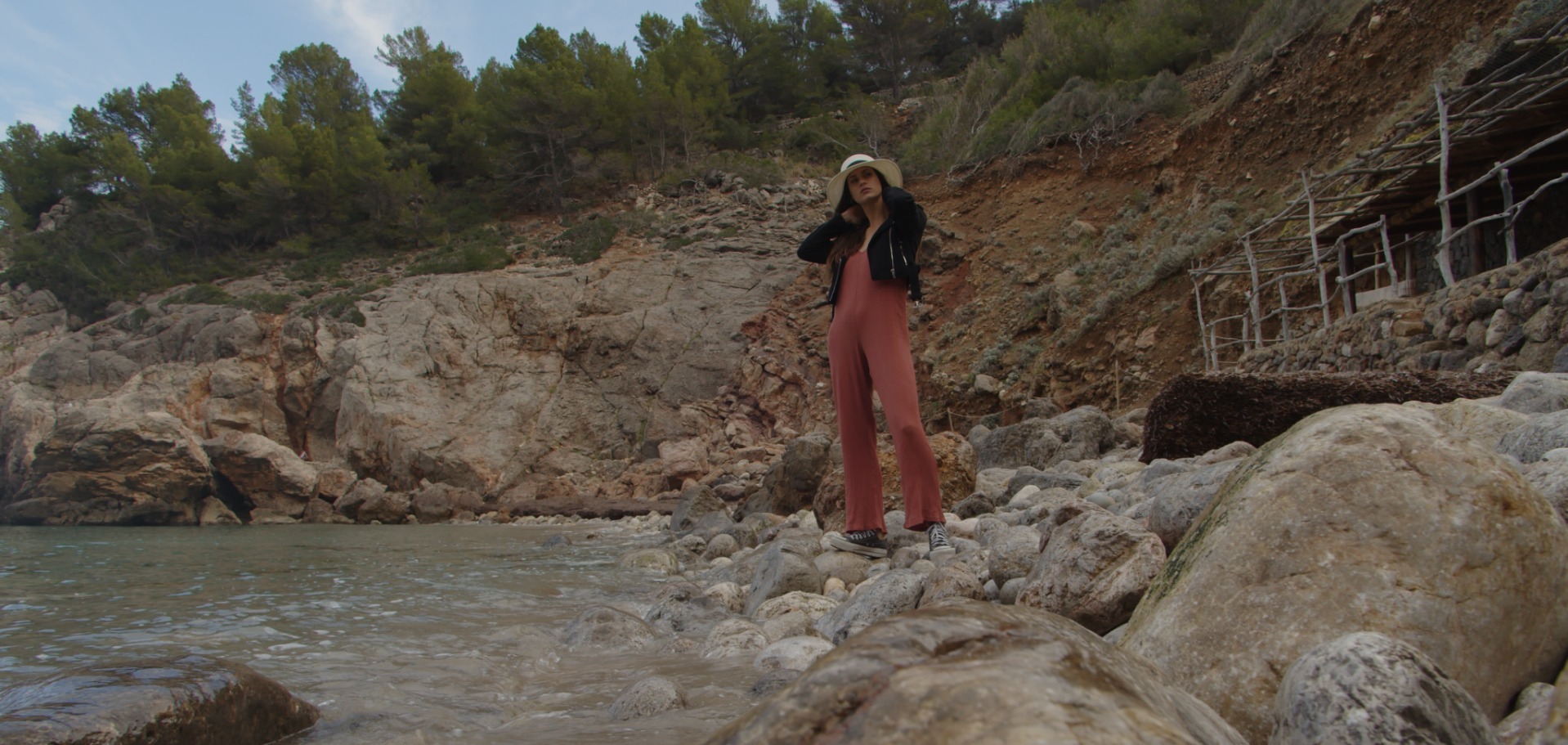 Kobieta w kombinezonie i kapeluszu pozuje na kamienistej plaży z klifami i morzem w tle.