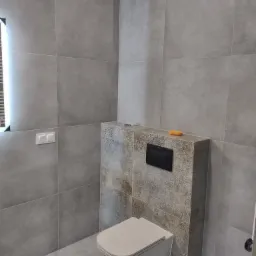 Nowoczesna łazienka z szarymi płytkami, wiszącą toaletą i dekoracyjną obudową stelaża z czarnym przyciskiem spłukującym, gąbka na obudowie.