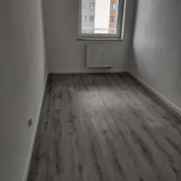 Pusty pokój z szarą podłogą imitującą drewno, świeżo pomalowanymi białymi ścianami, grzejnikiem pod oknem i widokiem na budynek mieszkalny za oknem.