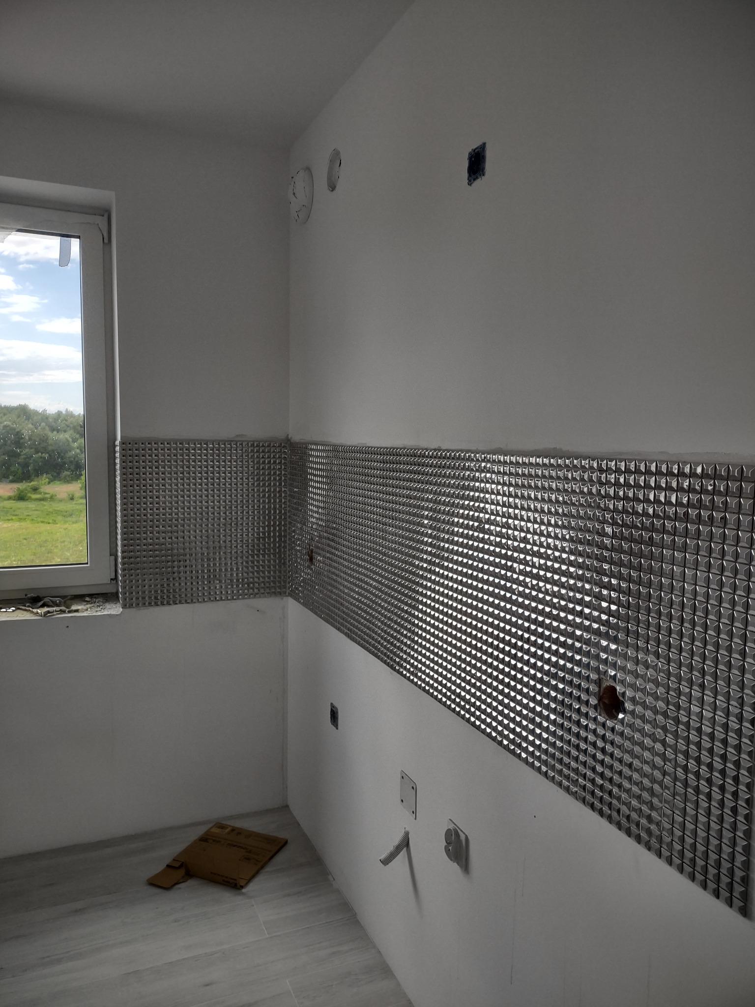 Montaż dekoracyjnych, srebrnych płytek mozaikowych na ścianie w kuchni, widoczne niedokończone otwory na instalacje elektryczne i hydrauliczne, okno z widokiem na zieleń, podłoga wyłożona szarymi...