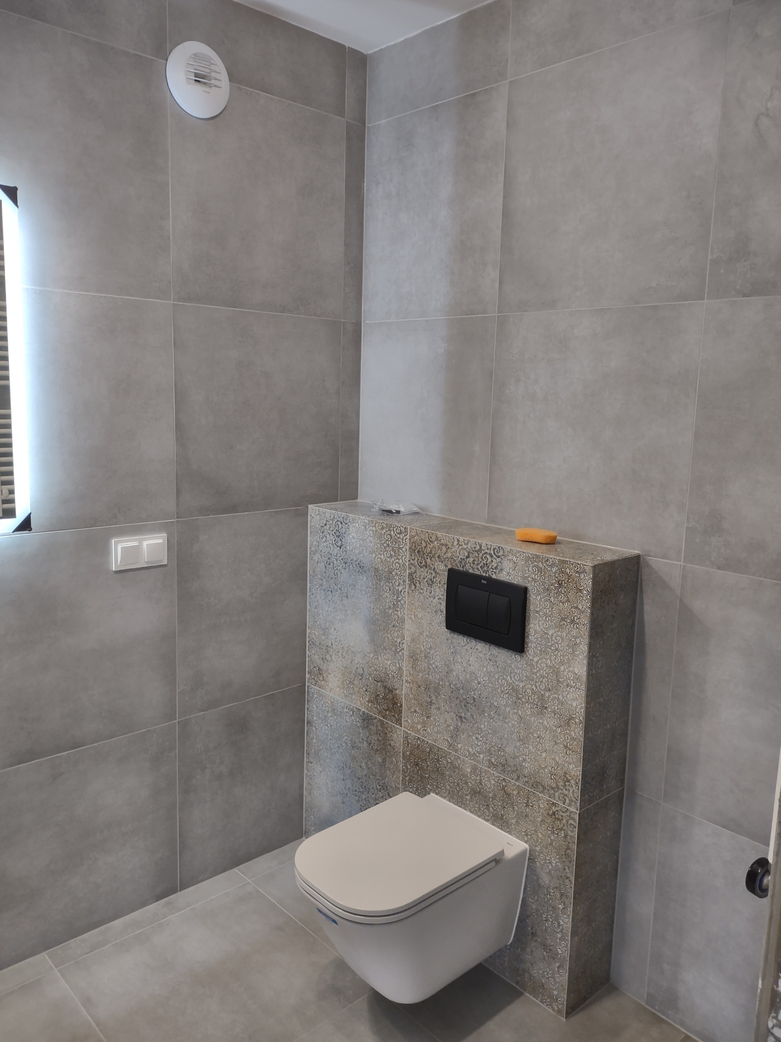 Nowoczesna łazienka z szarymi płytkami, wiszącą toaletą i dekoracyjną obudową stelaża z czarnym przyciskiem spłukującym, gąbka na obudowie.