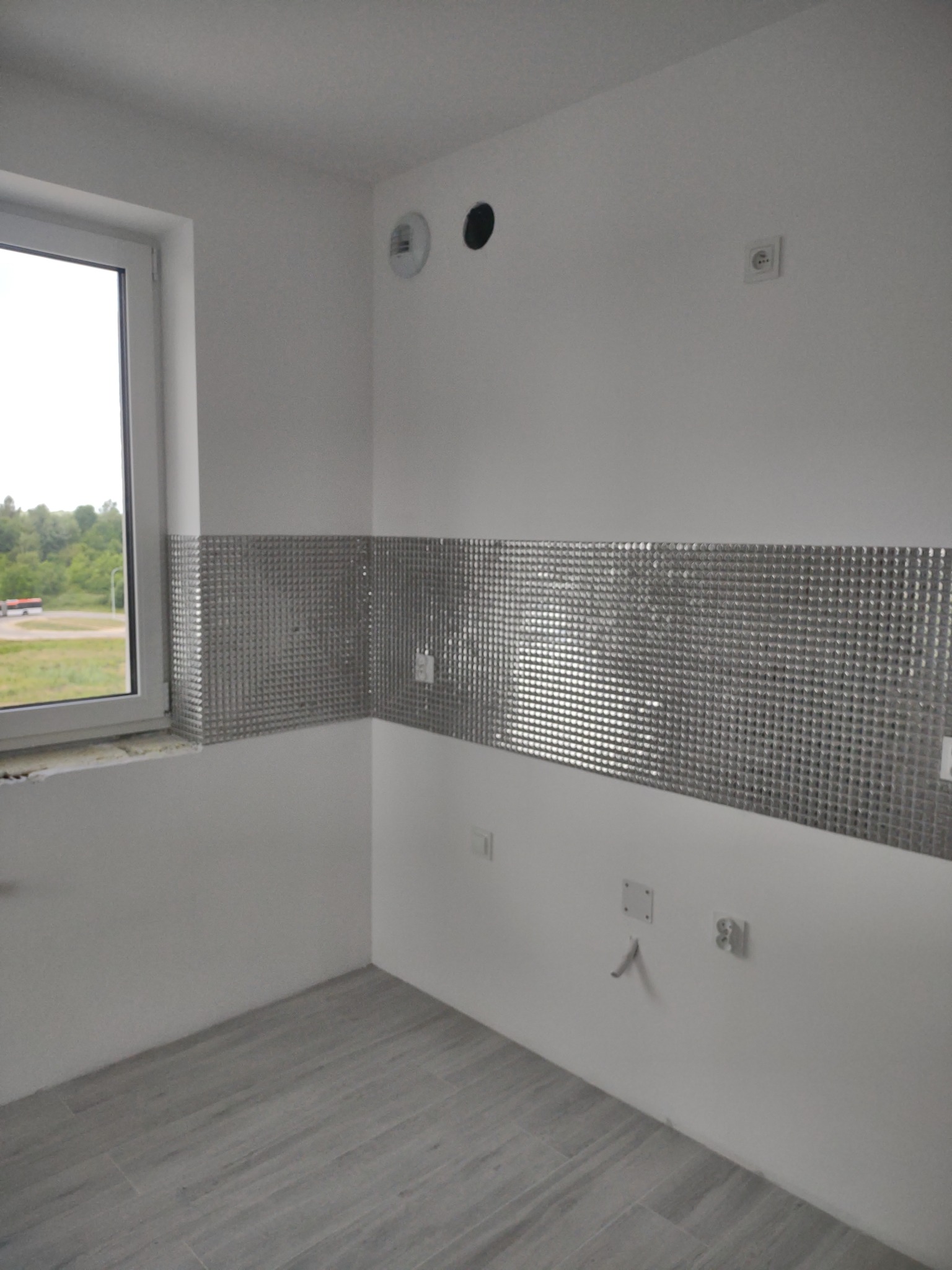 Fragment ściany w pomieszczeniu z częściowo ułożoną mozaiką w odcieniach szarości, widoczne gniazdka elektryczne i wyprowadzenie hydrauliczne, podłoga wyłożona panelami imitującymi drewno, okno...