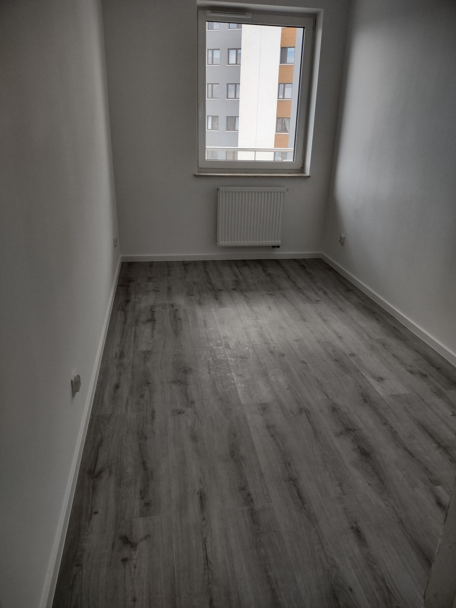Pusty pokój z szarą podłogą imitującą drewno, świeżo pomalowanymi białymi ścianami, grzejnikiem pod oknem i widokiem na budynek mieszkalny za oknem.