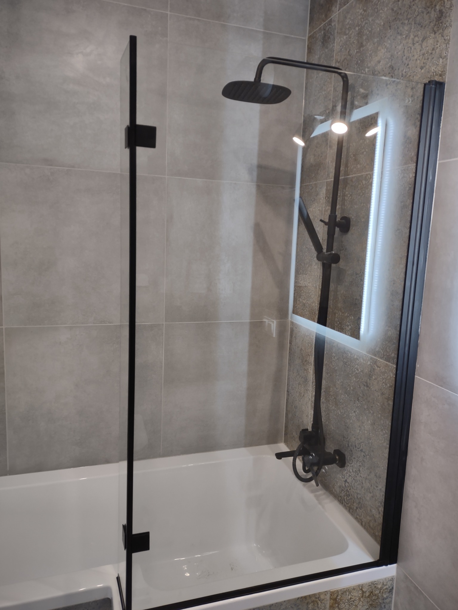 Nowoczesna łazienka z wanną i czarną armaturą: deszczownica, bateria, szklany parawan z czarnymi profilami, płytki w odcieniach szarości.