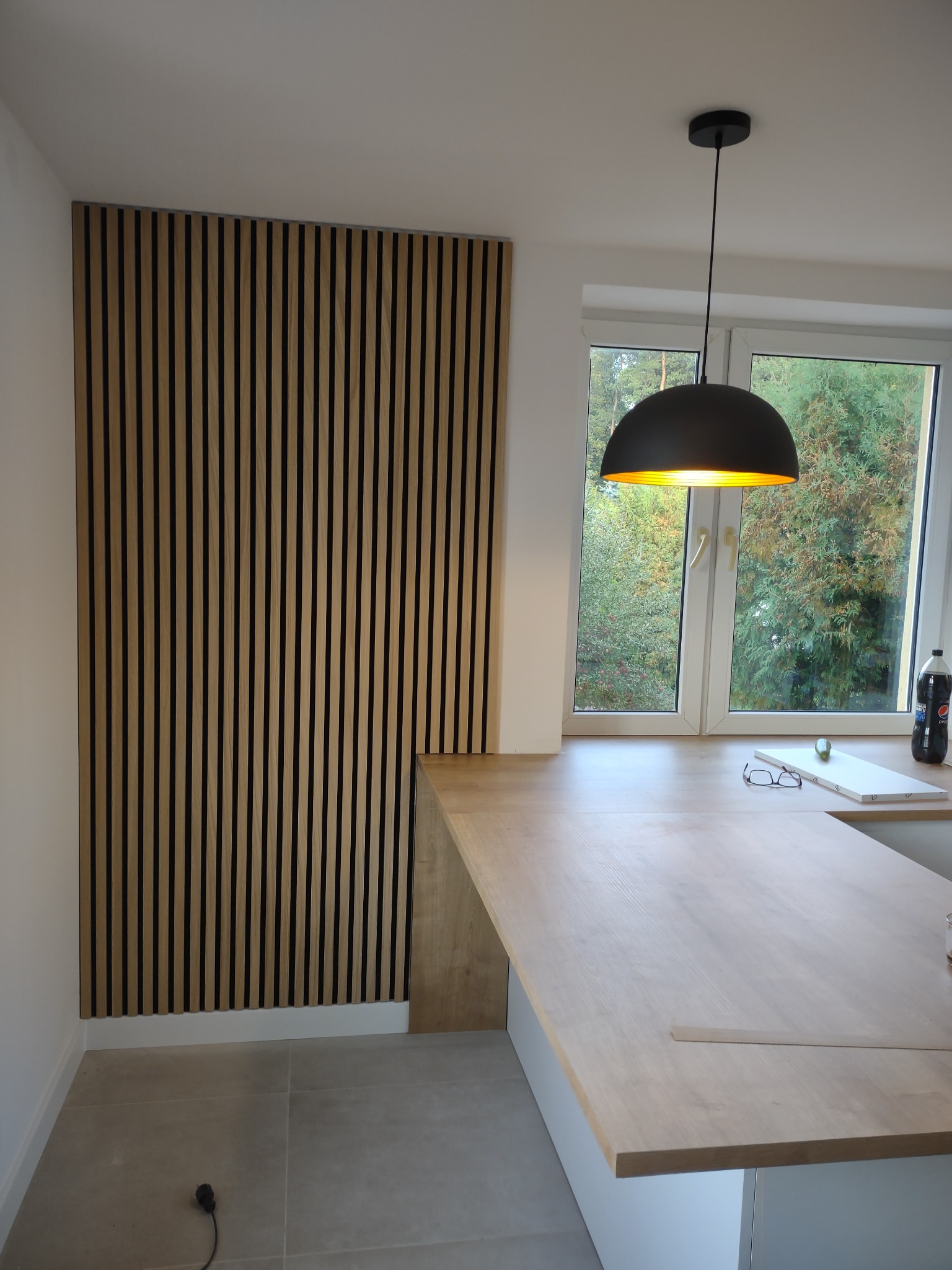Drewniane lamele na ścianie w jasnym wnętrzu, widoczna część blatu, lampa wisząca i okno z widokiem na zieleń.