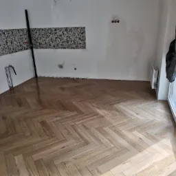 Nowo ułożona, jasna podłoga z paneli w jodełkę francuską w trakcie remontu; widoczne płytki dekoracyjne na ścianie, poziomica i zabezpieczona folią stolarka okienna.