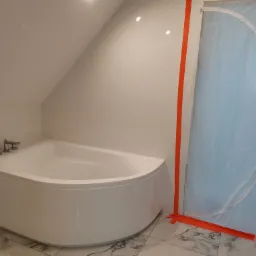 Biała, narożna wanna w łazience na poddaszu, zabezpieczona folią ochronną z pomarańczową taśmą malarską podczas remontu, widoczne baterie i marmurowe płytki podłogowe.