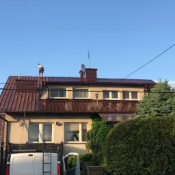 Pracownik na dachu dwupiętrowego domu z brązowym dachem, oparty o drabinę, z samochodem dostawczym poniżej, na tle błękitnego nieba.