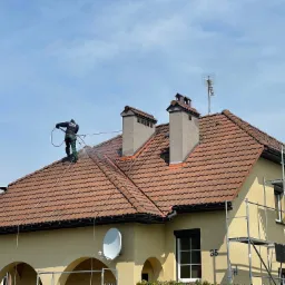 Pracownik w uprzęży, z użyciem myjki ciśnieniowej, czyści dach z ceramicznej dachówki, widoczne dwa kominy, antena telewizyjna oraz rusztowanie przy ścianie budynku.