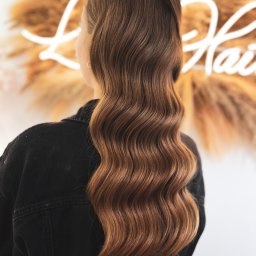 Oliwia Konopka - Brązowe, falowane włosy opadające na plecy kobiety ubranej w czarną kurtkę dżinsową. W tle dekoracja ze sztucznych traw i neonowy napis.