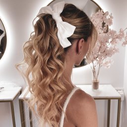 Oliwia Konopka - Upięcie włosów w Szczecinie: blond loki zebrane w wysoki kucyk, ozdobione dużą białą kokardą, widok z tyłu, delikatny makijaż, minimalistyczne kolczyki.