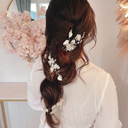 Oliwia Konopka - Upięcie włosów w warkocz z dekoracyjnymi, białymi spinkami w kształcie kwiatów i liści, widok z tyłu, na tle lustra i dekoracji kwiatowych.