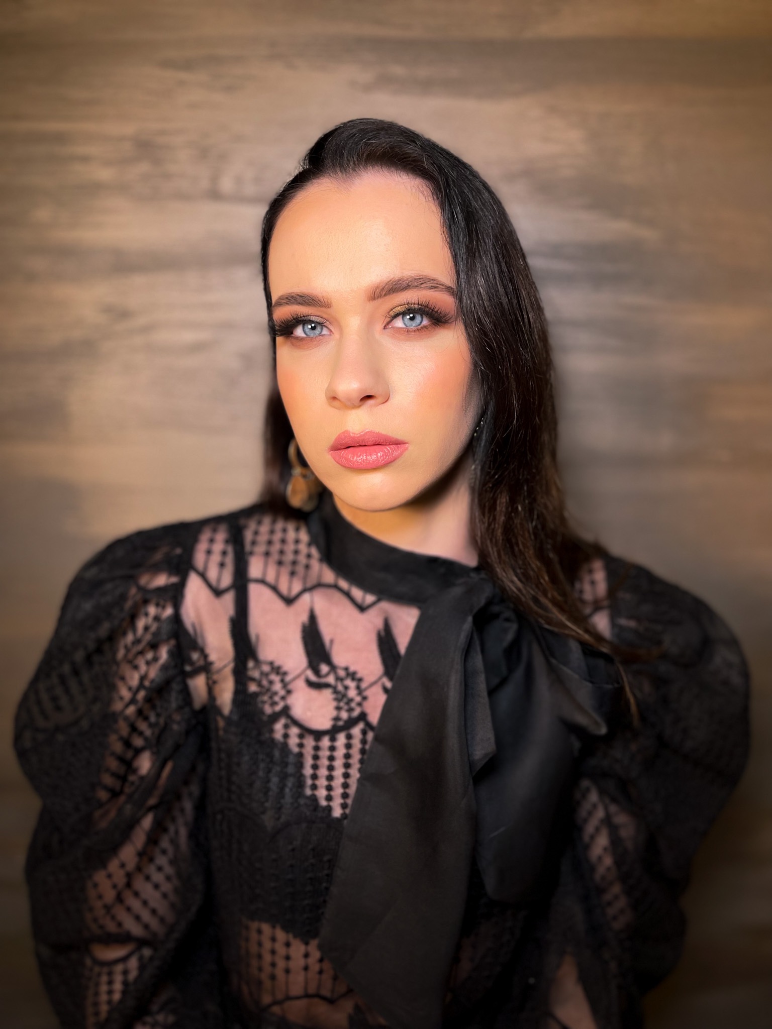 Portret kobiety z intensywnym makijażem oczu i ust w odcieniach nude, ubrana w czarną koronkową bluzkę z kokardą, na tle drewnianej ściany.