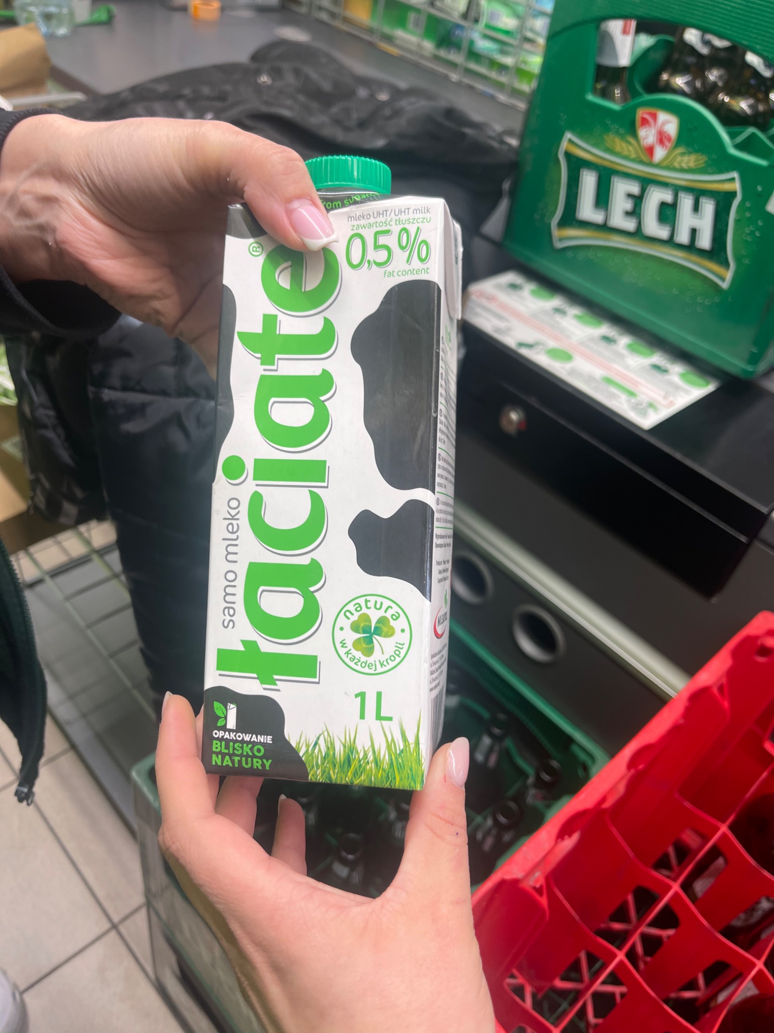 Dłoń z mlekiem 'Łaciate 0,5%' na tle sklepowym: zielony karton piwa Lech, czarna kurtka i czerwony koszyk. Paznokcie z delikatnym, jasnym, francuskim wzorem.